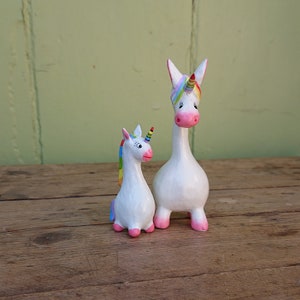 May include: Two white unicorn figurines with rainbow manes and pink hooves. The larger unicorn is standing upright, while the smaller unicorn is sitting down.