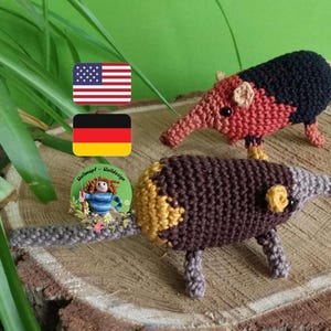 May include: Two crocheted stuffed animals shaped like anteaters. One is brown and yellow with a gray snout. The other is red, black, and orange with a brown snout. The animals are sitting on a wooden log.