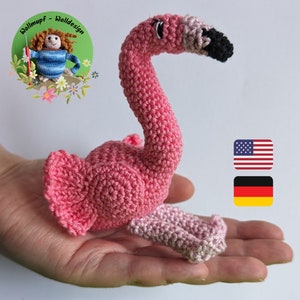 May include: Pink crocheted flamingo toy with black eyes and beak. The toy is held in a person's hand. The image also includes a small American flag and a German flag.