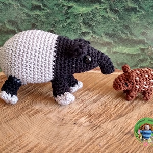 Könnte beinhalten: Zwei gehäkelte Stofftiere, ein graues und schwarzes Tapir und ein braunes und weißes Gürteltier, stehen auf einer Holzoberfläche. Das Tapir ist größer als das Gürteltier und hat eine lange Schnauze. Das Gürteltier hat einen kleinen, runden Körper und einen kurzen Schwanz.