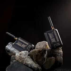 Könnte beinhalten: Zwei schwarze elektronische Geräte mit Antennen, beschriftet mit "COLD SWAP ONLY" und "SLEVIN DESIGN", liegen auf einem Steinhaufen vor dunklem Hintergrund. Die Geräte haben QR-Codes und USB-Anschlüsse.