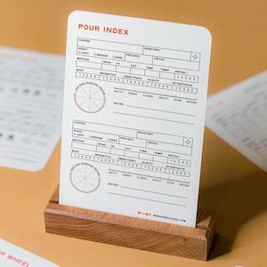 May include: A white card with a black and red "Pour Index" title and a circular chart with a scale of 1 to 5 for rating coffee characteristics like roast, grind, acidity, and aftertaste. The card is on a wooden stand.