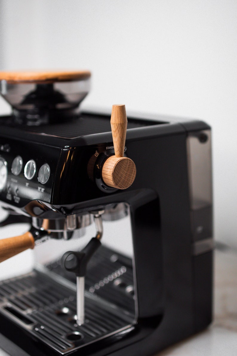 May include: A black espresso machine with a wooden handle on the steam wand. The machine has a stainless steel drip tray and a clear water tank.
