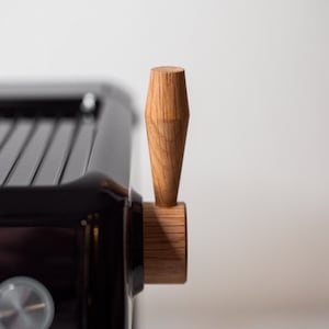 May include: Close-up of a black espresso machine with a wooden handle. The handle is a light brown color and has a slightly rounded shape.