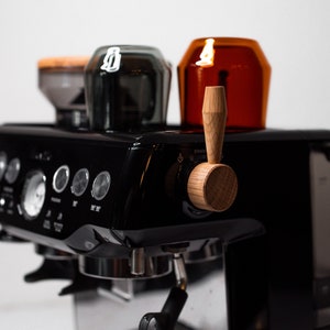 May include: A black espresso machine with a wooden lever and two portafilters. The machine has a digital display and several buttons. There are two glass containers on top of the machine.