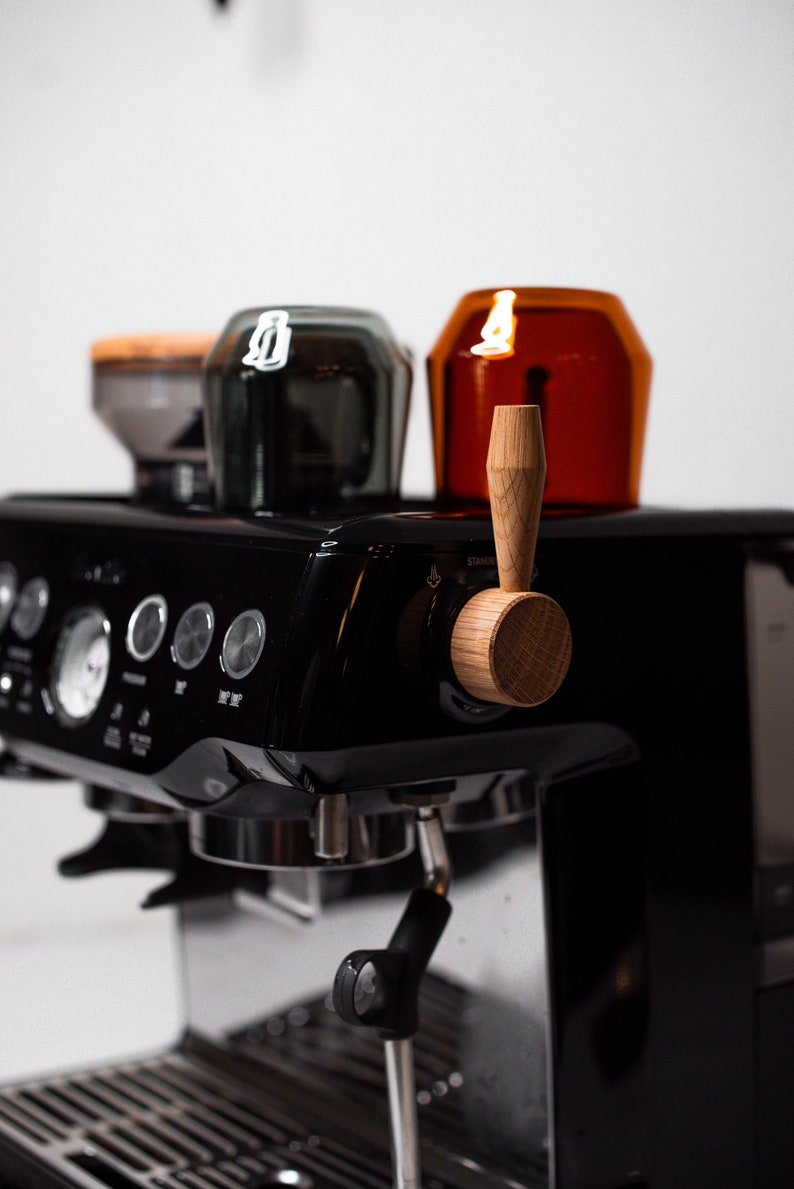 May include: A black espresso machine with a wooden lever and two portafilters. The machine has a digital display and several buttons. There are two glass containers on top of the machine.