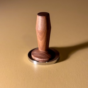 以下が含まれることがあります： 濃い茶色のハンドルと銀色のベースが付いた木製のコーヒータンパー。タンパーはコーヒー粉を均等に押すように設計されています。ハンドルは滑らかで人間工学に基づいた形状をしています。ベースは円形の研磨された金属です。