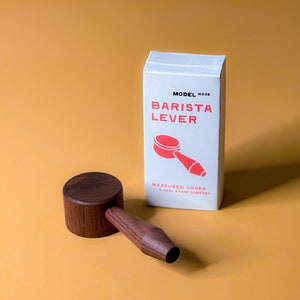 May include: A white box with the text "MODEL NOOB BARISTA LEVER" in red and a red illustration of a coffee tamper. The box is sitting on a yellow surface next to a wooden coffee tamper.