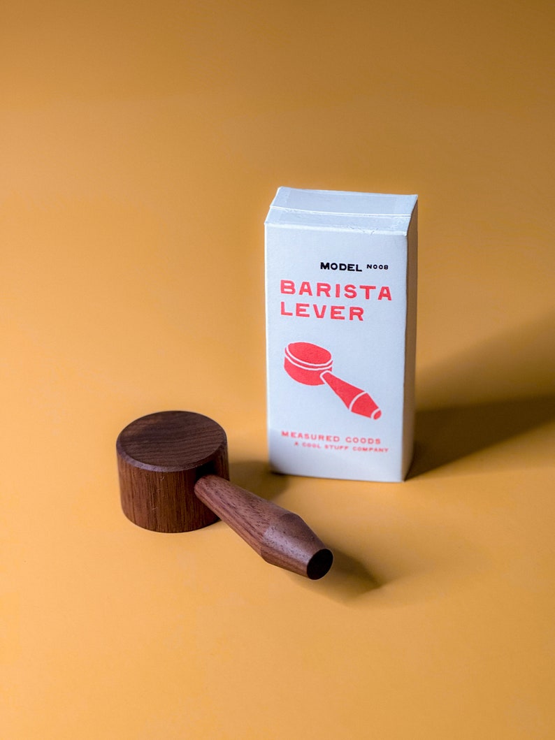 May include: A white box with the text "MODEL NOOB BARISTA LEVER" in red and a red illustration of a coffee tamper. The box is sitting on a yellow surface next to a wooden coffee tamper.