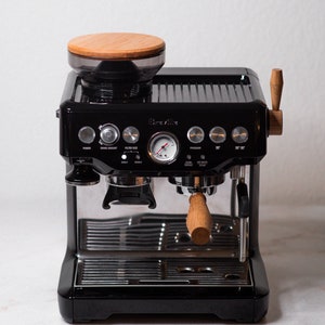 May include: A black Breville espresso machine with a wood-topped bean hopper, a stainless steel portafilter, and a wood handle. The machine has a variety of buttons and a pressure gauge.