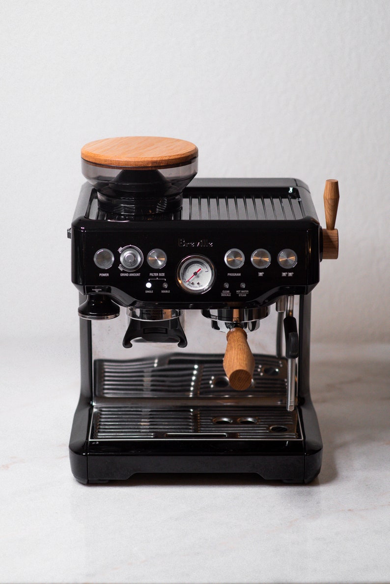 May include: A black Breville espresso machine with a wood-topped bean hopper, a stainless steel portafilter, and a wood handle. The machine has a variety of buttons and a pressure gauge.