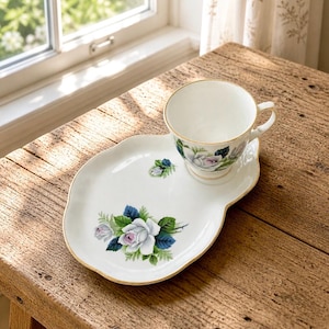 May include: A white teacup and saucer set featuring a rose design. The cup and saucer have a gold trim. The saucer is an irregular shape. The roses are white with pink centres and blue and green leaves. The set is on a wooden table.