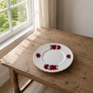 Puede incluir: Un plato de porcelana blanca con borde dorado y diseño floral de rosas rojas y hojas verdes. El plato está sobre una mesa de madera rústica cerca de una ventana.