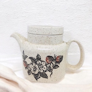May include: A white ceramic teapot with a speckled glaze and a floral design featuring black leaves, white flowers, and red berries. The teapot has a lid and a handle.