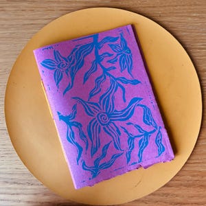 Peut inclure: Un petit carnet violet avec un motif floral bleu, posé sur une surface ronde jaune-orangé. La couverture du carnet présente un motif répété de fleurs et de feuilles stylisées. Le fond est une surface en bois.