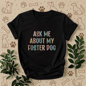 Peut inclure: T-shirt noir avec l'inscription "ASK ME ABOUT MY FOSTER DOG" en couleurs pastel. Le t-shirt a un col rond et des manches courtes. Le fond présente des illustrations de chiens et de chats, des empreintes de pattes et des os.