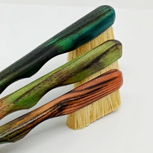 May include: Three wooden handled brushes with natural bristles. The handles are stained in shades of green and red-brown. The bristles are a light tan color. The brushes are arranged diagonally against a white background.