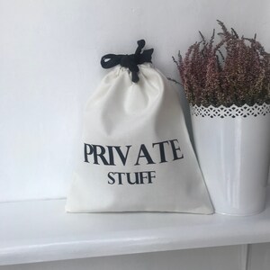 Peut inclure: Un sac en toile blanc avec des cordons de serrage noirs et le texte "PRIVATE STUFF" imprimé en noir.