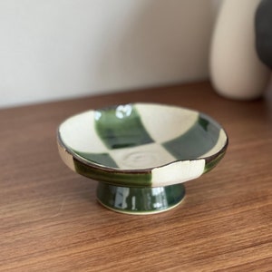 May include: A small, ceramic pedestal bowl with a green and cream checkerboard pattern. The bowl has a rounded shape and a short, cylindrical base. The rim is a dark brown colour. The bowl is displayed on a wooden surface.