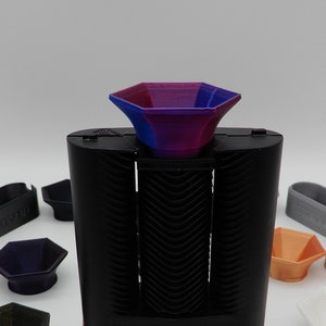 Könnte beinhalten: Ein schwarzer elektronischer Vaporizer mit einem digitalen Display und orangefarbenen Akzenten. Der Vaporizer hat ein lila-blaues Mundstück und ist von verschiedenen farbigen, sechseckigen Accessoires umgeben.