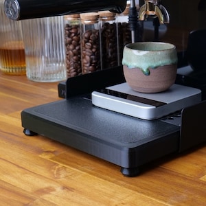 Könnte beinhalten: Eine schwarze Espressomaschine mit einer Keramiktasse auf einer digitalen Waage. Die Tasse ist braun mit einer grünen Glasur. Gläser mit Kaffeebohnen und Flüssigkeiten sind im Hintergrund. Die Maschine steht auf einer Holzoberfläche.