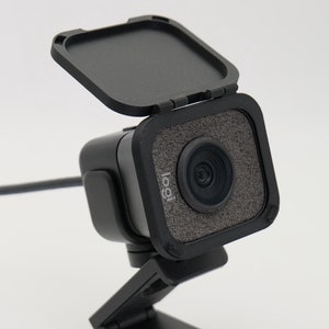 Könnte beinhalten: Eine schwarze Webcam mit einem geöffneten Datenschutzverschluss, der ein schwarzes Objektiv freigibt. Die Webcam hat eine strukturierte schwarze Oberfläche und einen kleinen, verstellbaren Ständer.