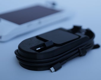 Może przedstawiać: Czarna stacja dokująca z kablem USB-C owiniętym wokół niej. Stacja dokująca ma mały, prostokątny otwór, w który można umieścić urządzenie.