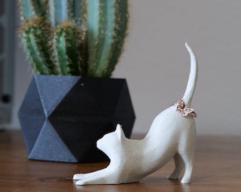Peut inclure: Une figurine de chat blanc en position d'étirement, avec une bague papillon en or rose sur sa queue. Un cactus vert est dans un pot géométrique noir en arrière-plan. La figurine de chat est sur une surface en bois.