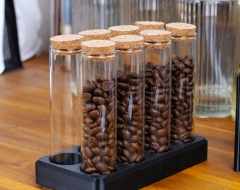 Puede incluir: Un conjunto de tubos de vidrio transparente con tapones de corcho, llenos de granos de café, presentados en un soporte negro. Los tubos están dispuestos sobre una superficie de madera, con otros recipientes de vidrio en el fondo. También se ve un soporte negro.