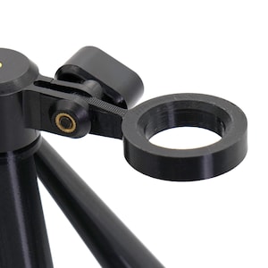 May include: Close-up of a black tripod head with a circular ring attachment. The tripod head has a level indicator and a hinged arm with a gold-colored screw. The tripod legs are black and angled.