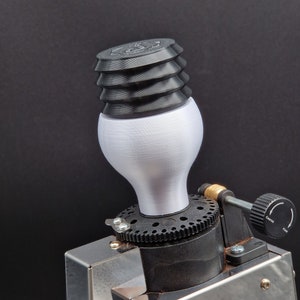 Könnte beinhalten: Nahaufnahme einer silbernen Kaffeemühle mit einem schwarzen und weißen Mahlgrad-Einstellknopf. Die Mühle hat ein schwarzes Plastik-Oberteil mit einem weißen, runden, strukturierten Mahlgutbehälter.