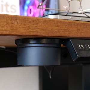 Peut inclure: Gros plan sur un panneau de commande noir pour le réglage de la hauteur d'un bureau, avec des boutons M, 1, 2, 3 et 4. Un objet cylindrique noir est monté sous un bureau en bois. Des lunettes rondes et un téléphone sont posés sur le bureau.