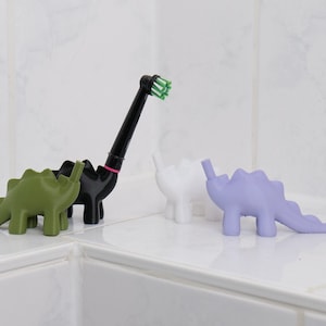 Könnte beinhalten: Vier dinosaurierförmige Zahnbürstenhalter in Grün, Schwarz, Weiß und Lila. Einer der Halter hat eine schwarze elektrische Zahnbürste mit einem grünen Bürstenkopf, der in ihn eingesetzt ist.