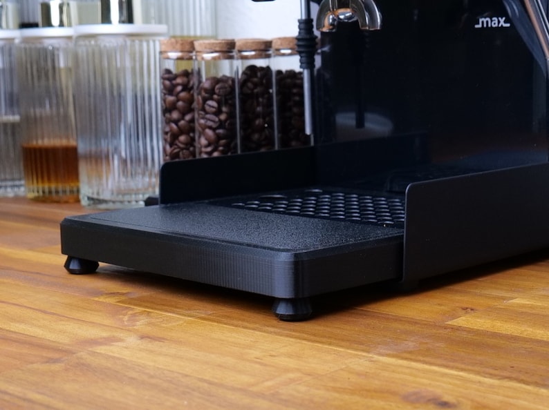 May include: Black espresso machine with a textured base and small, rounded feet. The machine has a drip tray and is set on a wooden surface. Glass jars with coffee beans are visible in the background.