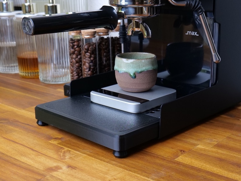 May include: A black espresso machine with a silver portafilter and a cup on a scale. The machine has a black drip tray and a black base with rubber feet. The cup is a brown ceramic mug with a green glaze.