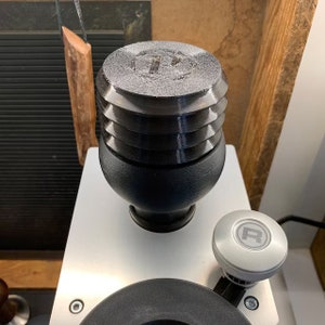 Könnte beinhalten: Eine schwarze und silberne Kaffeemühle mit einer runden, schwarzen, strukturierten Oberseite. Die Mühle hat eine silberne Basis und einen silbernen Knopf mit dem Buchstaben "R" darauf.