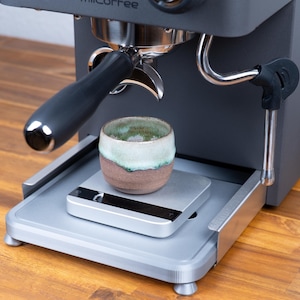 May include: A dark grey espresso machine with a portafilter and a small ceramic cup. The cup is brown with a green and white rim, sitting on a silver scale. The machine is on a wooden surface.