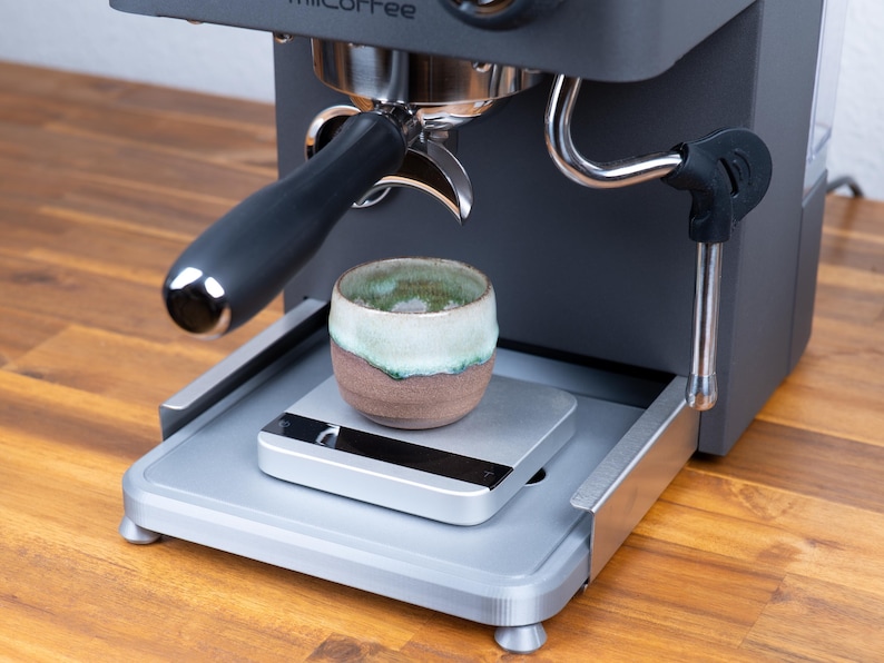 May include: A dark grey espresso machine with a portafilter and a small ceramic cup. The cup is brown with a green and white rim, sitting on a silver scale. The machine is on a wooden surface.
