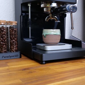May include: A black espresso machine with a ceramic cup on a scale. Glass tubes filled with coffee beans sit in a holder. The holder has the text "Paralyse3D". The cup has a green and brown glaze.