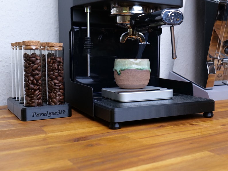 May include: A black espresso machine with a ceramic cup on a scale. Glass tubes filled with coffee beans sit in a holder. The holder has the text "Paralyse3D". The cup has a green and brown glaze.