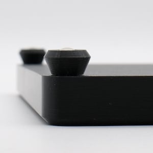 May include: Close-up of a black, rectangular object with two small, dark, conical feet. The object has a textured surface and a beveled edge. The background is plain white.