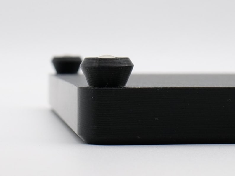 May include: Close-up of a black, rectangular object with two small, dark, conical feet. The object has a textured surface and a beveled edge. The background is plain white.