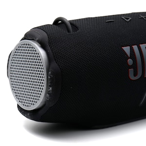 Könnte beinhalten: Schwarzer tragbarer JBL-Lautsprecher mit zylindrischem Design. Der Lautsprecher hat eine Mesh-Lautsprecherabdeckung an einem Ende und das JBL-Logo in Rot und Grau. Er verfügt über Bedientasten und einen Tragegriff.