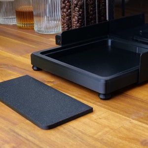 May include: A black coffee station organiser with a rectangular base and a separate textured mat. The organiser has a tray and raised sides. The items are on a wooden surface, with coffee beans and glasses in the background.