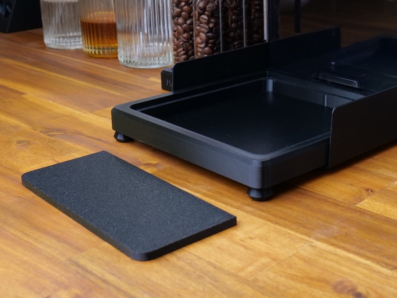 May include: A black coffee station organiser with a rectangular base and a separate textured mat. The organiser has a tray and raised sides. The items are on a wooden surface, with coffee beans and glasses in the background.
