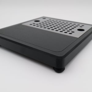May include: A black square product with a perforated metal section. The product has rounded corners and sits on four small, round feet. The top surface is matte black, and the perforated section is silver.