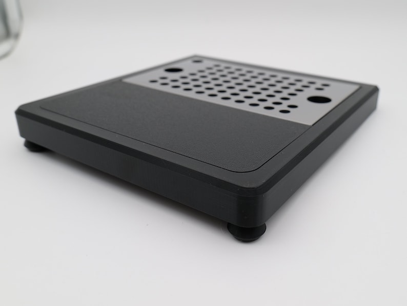 May include: A black square product with a perforated metal section. The product has rounded corners and sits on four small, round feet. The top surface is matte black, and the perforated section is silver.
