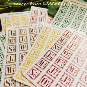 Peut inclure: Plusieurs feuilles d'autocollants avec des abréviations de calendrier mensuel de différentes couleurs, dont le rouge, l'or et le vert. Chaque autocollant présente une abréviation de mois dans un cadre carré. Le site web www.enviedecreer.fr est visible.
