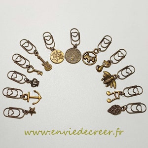 Könnte beinhalten: Ein Set mit 12 goldfarbenen Lesezeichenclips aus Metall mit verschiedenen Anhängern, darunter eine Gitarre, ein Baum, eine Katze, eine Biene, eine Musiknote und ein Anker. Die Anhänger sind mit einer kleinen Schlaufe an den Clips befestigt.