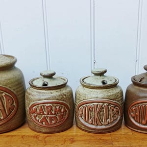 May include: A set of four ceramic kitchen canisters with lids, featuring speckled beige and brown glaze. Each canister has a raised oval emblem with embossed text reading SUGAR, MARMALADE, PICKLES, and CHUTNEY in a reddish-brown tone.